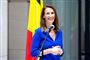 En smilende kvinde i blå bluse foran en mikrofon med det belgiske flag i baggrunden