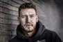 Nærbillede af fodboldspilleren Nicklas Bendtner. Han har et lille fuldskæg og et dybt alvorligt blik. Han er iført mørk hættetrøje og står op ad en murstens mur. 
