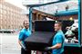 To smilende yngre mennesker slæber en sofa ind i en lastbil
