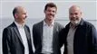 Tre mænd står tæt sammen med havbugten som baggrund