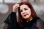 Priscilla Presley iklædt sort kjole og har den ene hånd oppe ved sit meget mørkrøde hår
