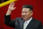 Kim Jong-un har sort jakkesæt og hvid skjorte og slips på. Står bag en mikrofon og har den ene arm i vejret som til en hilsen