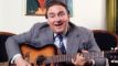 mand sidde i stol og synger med guitar
